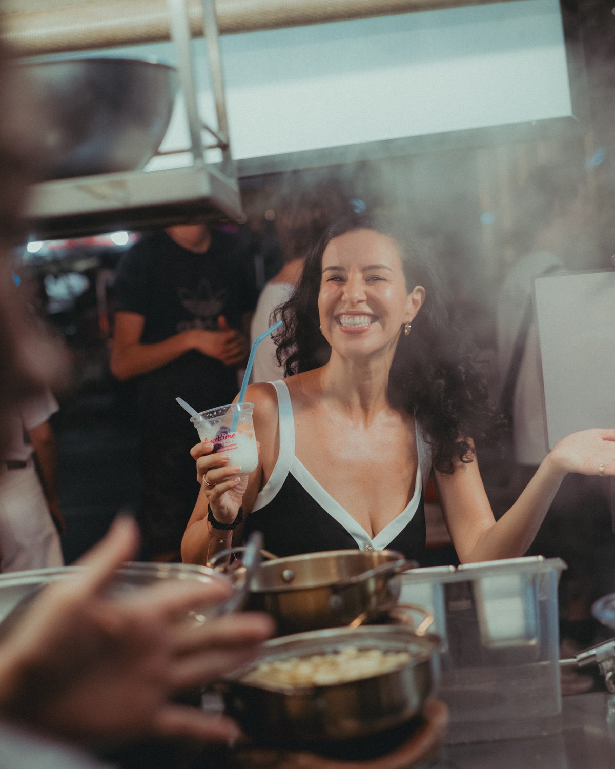 Amelle éclate de rire, un dessert glacé à la main, devant un stand de street food à Bangkok.